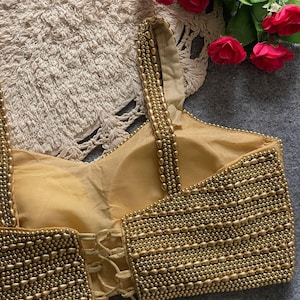 May include: A gold-coloured, beaded top with a corset-style back. The top features rows of small, round beads and a lace-up closure. The straps are also adorned with beads. Red roses are visible in the background.