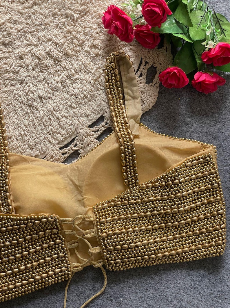 May include: A gold-coloured, beaded top with a corset-style back. The top features rows of small, round beads and a lace-up closure. The straps are also adorned with beads. Red roses are visible in the background.
