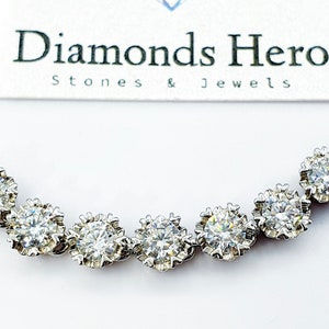 Op de afbeelding: Een zilveren ketting met een rij van zeven ronde, glinsterende, witte edelstenen die in een clusterdesign zijn gezet. De ketting ligt op een witte achtergrond met een kaart waarop "Diamonds Hero Stones & Jewels" staat.