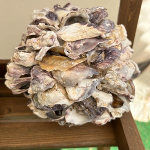 May include: A spherical decorative object constructed from numerous overlapping oyster shells. The shells display a natural color palette of off-white, tan, and deep purple-grey tones, creating a textured, layered surface across the entire round form.