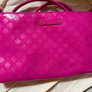 May include: A bright pink patent leather handbag featuring a repeating embossed spade pattern across the surface. The bag includes two top handles and a rectangular metal plaque centered at the top with the text Kate Spade New York engraved.