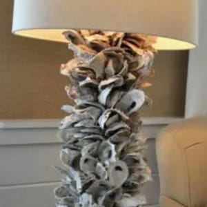 May include: A table lamp featuring a cylindrical base covered in layered, natural gray and white oyster shells. The lamp stands on a square, distressed white wooden base and is topped with a large, cylindrical, off-white fabric lampshade.