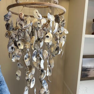 May include: A hanging mobile featuring two stacked circular wooden hoops from which numerous natural oyster shells are suspended by thin white strings. The shells display varied shades of gray, brown, tan, and white, creating a cascading effect.