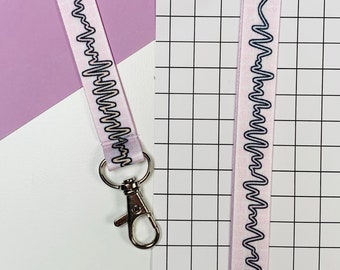May include: A pink lanyard with a black and white wavy line design and a silver clip. The lanyard is on a pink and white background.