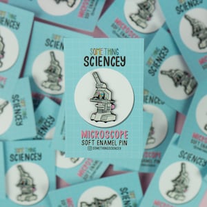 Puede incluir: Un pin de esmalte suave de microscopio blanco con detalles rosas sobre un fondo azul. El pin está rodeado de otros pines y tarjetas con el texto "Something Sciencey" en ellos.