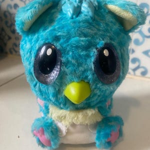 May include: A turquoise plush toy with large, reflective eyes and a yellow beak. The toy has a fluffy texture, with pink accents on its paws and ears. The toy is designed to resemble a bird or a similar creature.