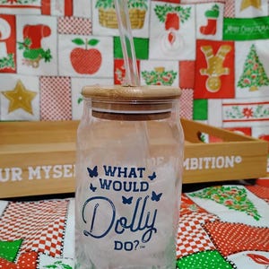 May include: A clear glass tumbler with a bamboo lid and a clear straw. The tumbler features the phrase "WHAT WOULD Dolly DO?" in blue script with butterfly accents. The background includes a Christmas-themed fabric.