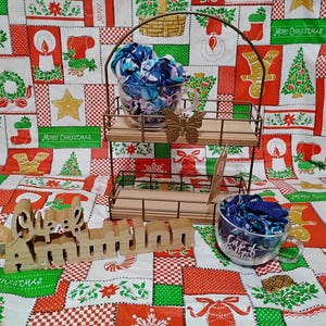 Könnte beinhalten: Ein zweistufiger Displayständer aus Metall und Holz hält zwei klare Teetassen aus Glas, die mit blauem Stoff gefüllt sind. Ein Holzschild mit der Aufschrift "Animal" befindet sich vor dem Ständer. Der Hintergrund ist eine weihnachtliche Tischdecke.