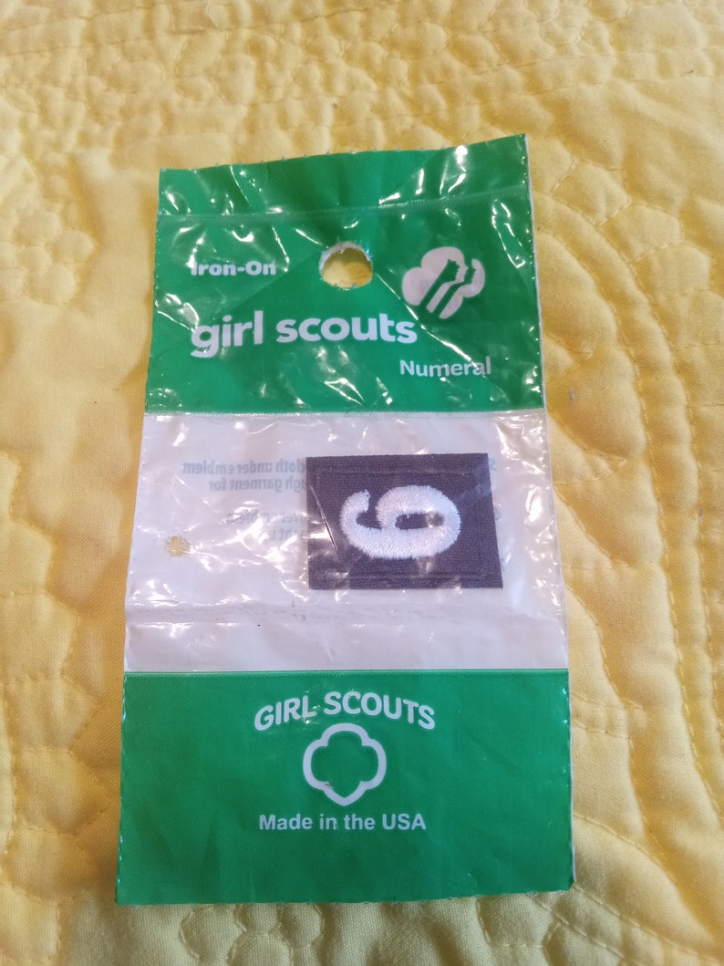 May include: A plastic package containing an iron-on Girl Scout numeral patch. The patch is gray with a white number 6. The package is green and white with the text "Iron-On Girl Scouts Numeral" and "Girl Scouts Made in the USA".
