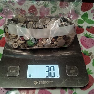 May include: A digital kitchen scale with a white display showing 3.0, a white button labeled "UNIT", and a white button labeled "ZERO". A clear plastic bag filled with a variety of buttons is on the scale.