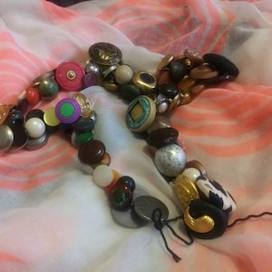 May include: A colorful necklace made of various buttons, beads, and other trinkets. The necklace is laid out on a white and pink patterned fabric.