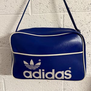 Może przedstawiać: Niebieska torba na ramię z białym wykończeniem i białym logo Adidas. Torba ma prostokątny kształt z długim paskiem. Logo i tekst Adidas są widoczne z przodu. Szerokość: około 30 cm.