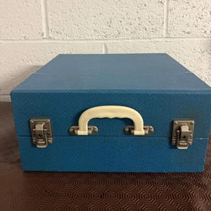 Op de afbeelding: Een vierkante, blauwe, getextureerde opbergkoffer met een crèmekleurig kunststof handvat in het midden van het voorpaneel. De koffer wordt afgesloten met twee zilverkleurige metalen sluitingen aan weerszijden van het handvat voor stevigheid.
