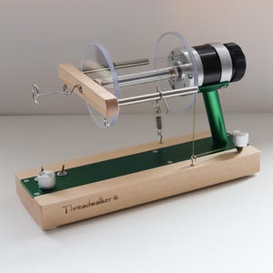Puede incluir: Un devanador de hilo de madera y metal, con un acento verde. Tiene una bobina de plástico transparente y una base de madera con el texto "Threadwalker". Diseñado para enrollar hilo en una madeja.