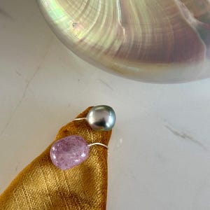 Può includere: Due anelli con fasce argentate, uno con una perla grigia e l'altro con una gemma rosa, poggiano su un panno dorato. Sullo sfondo, una grande conchiglia di nautilus iridescente riflette la luce con sfumature di bianco, giallo e blu.