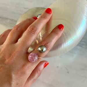 Op de afbeelding: Een goudkleurige ring met een roze, gebarsten glazen bol en een grijze parel. De ring zit aan een hand met rode nagellak, tegen een witte en iriserende achtergrond.