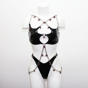 May include: Black vinyl lingerie set with silver chain accents and red spider charms. The set includes a top, a thong, and connecting chains. The lingerie is displayed on a white mannequin against a white background.