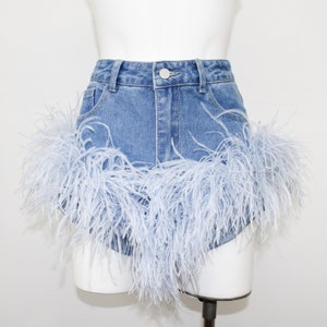 May include: Light blue denim shorts with a high waist and a single button closure. The shorts are trimmed with a fluffy, light blue feather detail around the hemline. The shorts are displayed on a white mannequin.