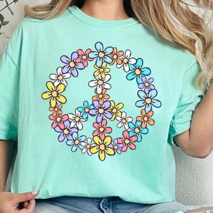 以下が含まれることがあります： カラフルな花でできた平和のサインが描かれた薄い青色のTシャツ。平和のサインはTシャツの中央にあります。