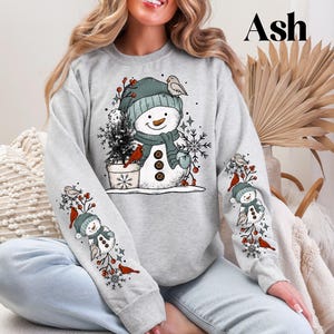 K&ouml;nnte beinhalten: Graues Sweatshirt mit einem Schneemann-Design, gr&uuml;ner M&uuml;tze, Schal und Kn&ouml;pfen. Die &Auml;rmel haben passende Schneemann- und Kardinalvogel-Illustrationen. Ein kleiner Weihnachtsbaum befindet sich neben dem Schneemann. Das Wort "Ash" ist oben rechts aufgedruckt.