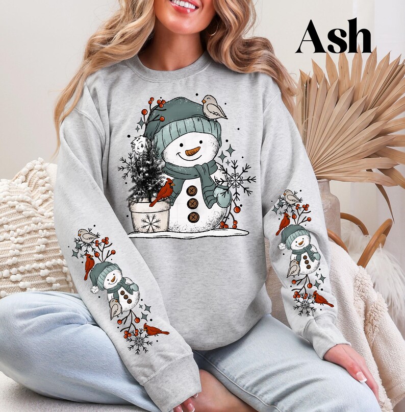 K&ouml;nnte beinhalten: Graues Sweatshirt mit einem Schneemann-Design, gr&uuml;ner M&uuml;tze, Schal und Kn&ouml;pfen. Die &Auml;rmel haben passende Schneemann- und Kardinalvogel-Illustrationen. Ein kleiner Weihnachtsbaum befindet sich neben dem Schneemann. Das Wort "Ash" ist oben rechts aufgedruckt.