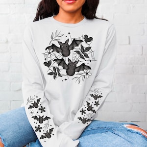 K&ouml;nnte beinhalten: Wei&szlig;es Langarmshirt mit einem schwarzen Fledermaus- und Blumendesign auf der Vorderseite und schwarzen Flederm&auml;usen auf den &Auml;rmeln.