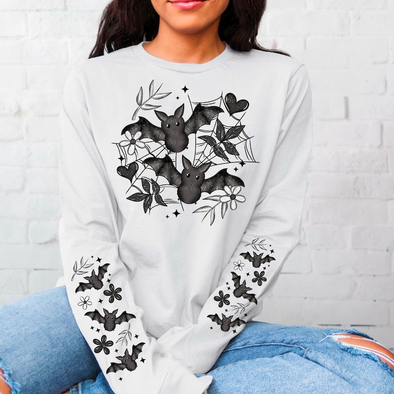 K&ouml;nnte beinhalten: Wei&szlig;es Langarmshirt mit einem schwarzen Fledermaus- und Blumendesign auf der Vorderseite und schwarzen Flederm&auml;usen auf den &Auml;rmeln.