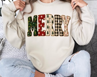 May include: A cream-coloured sweatshirt featuring the word "MERRY" in a festive design. The letters are decorated with patterns including holly, stars, Christmas trees, plaid, and berries. A casual, holiday-themed garment.