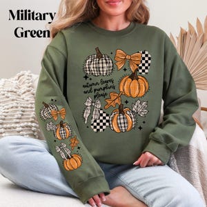 May include: Military green sweatshirt with a fall-themed design. The design includes orange pumpkins, plaid bows, and the text "autumn leaves and pumpkins please". The sweatshirt is a comfortable, casual style.