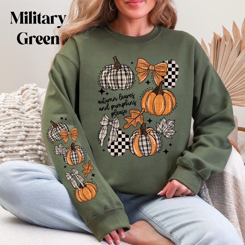 May include: Military green sweatshirt with a fall-themed design. The design includes orange pumpkins, plaid bows, and the text "autumn leaves and pumpkins please". The sweatshirt is a comfortable, casual style.
