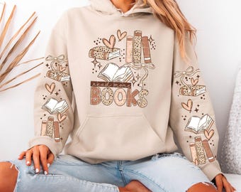 May include: Beige hoodie with a book-themed graphic. The design features open books, stacks of books, hearts, and the text "read more BOOKS". The sleeves also have book and heart designs. A cozy and stylish sweatshirt for book lovers.
