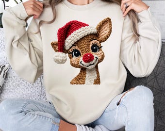 May include: A cream-colored sweatshirt featuring a detailed graphic of a reindeer wearing a Santa hat. The reindeer has a red nose, large eyes, and a smiling expression. The sweatshirt is a festive holiday design.