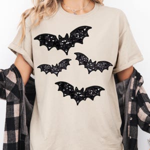 May include: Beige t-shirt featuring four black sequined bat designs. The bats are arranged in a scattered pattern across the front of the shirt. The shirt has short sleeves and a relaxed fit, suitable for casual wear.