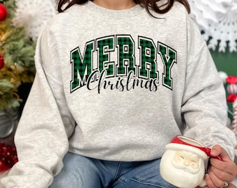 Op de afbeelding: Een lichtgrijze sweatshirt met de tekst "MERRY Christmas". Het woord "MERRY" is in een groen en zwart geruit ontwerp, in een vetgedrukte blokletter. "Christmas" is in een cursief lettertype. Een feestelijk, casual kledingstuk voor de feestdagen.
