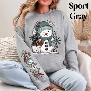 K&ouml;nnte beinhalten: Graues Sport Gray Sweatshirt mit einem Schneemann-Design, das einen Hut, Schal und F&auml;ustlinge tr&auml;gt. Der Schneemann ist mit einem Vogel, Schneeflocken und einem kleinen Weihnachtsbaum verziert. Der &Auml;rmel hat ein passendes Design. Der Text "Sport Gray" befindet sich oben rechts.