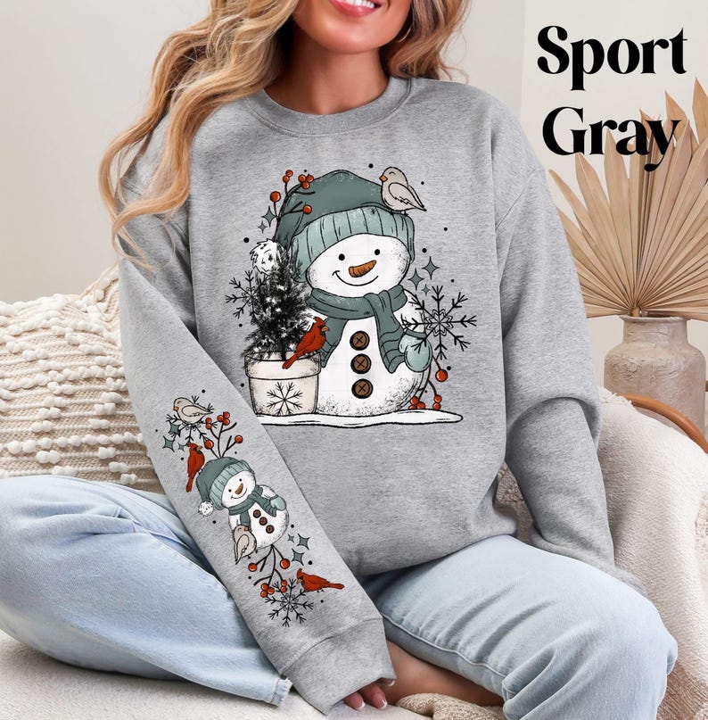 K&ouml;nnte beinhalten: Graues Sport Gray Sweatshirt mit einem Schneemann-Design, das einen Hut, Schal und F&auml;ustlinge tr&auml;gt. Der Schneemann ist mit einem Vogel, Schneeflocken und einem kleinen Weihnachtsbaum verziert. Der &Auml;rmel hat ein passendes Design. Der Text "Sport Gray" befindet sich oben rechts.