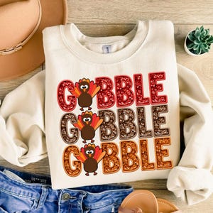Puede incluir: Una sudadera de color crema con las palabras "Gobble Gobble Gobble" impresas en purpurina roja, marr&oacute;n y naranja. Cada palabra tiene un pavo de dibujos animados con plumas marrones y car&uacute;nculas rojas.