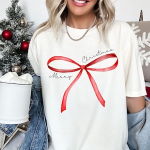 May include: Cream-colored t-shirt featuring a watercolor-style red bow design. The words "Merry Christmas" are written in black script. The shirt is a casual, festive apparel item, perfect for the holiday season.