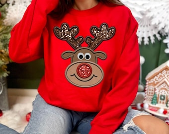 May include: A red sweatshirt featuring a reindeer design. The reindeer has brown antlers with sequin accents, black eyes, and a red sequined nose. The sweatshirt is a solid red color.
