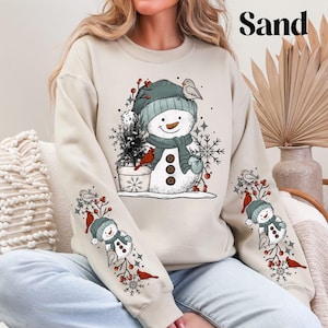 K&ouml;nnte beinhalten: Sandfarbenes Sweatshirt mit einem Schneemann-Design. Der Schneemann tr&auml;gt einen blauen Hut und Schal, eine Karottennase und Knopfaugen. Die &Auml;rmel zeigen kleinere Schneemann- und Vogelillustrationen. Das Wort "Sand" ist oben aufgedruckt.