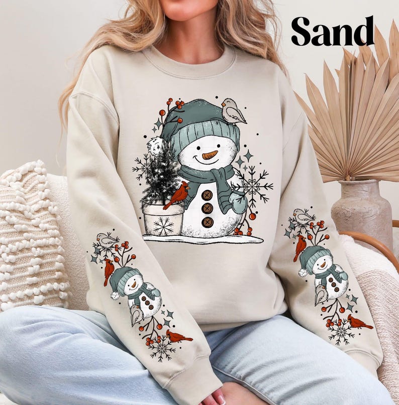 K&ouml;nnte beinhalten: Sandfarbenes Sweatshirt mit einem Schneemann-Design. Der Schneemann tr&auml;gt einen blauen Hut und Schal, eine Karottennase und Knopfaugen. Die &Auml;rmel zeigen kleinere Schneemann- und Vogelillustrationen. Das Wort "Sand" ist oben aufgedruckt.