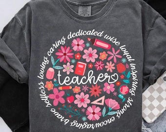 May include: A gray t-shirt with a white circle design featuring pink flowers, school supplies, and the word "teacher" surrounded by the words "strong, inspiring, loyal, wise, dedicated, caring, loving, selfless, brave, encouraging".
