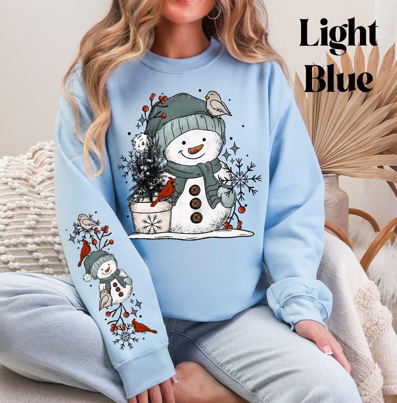 K&ouml;nnte beinhalten: Hellblaues Sweatshirt mit einem Schneemann-Design. Der Schneemann tr&auml;gt einen gr&uuml;nen Hut, Schal und F&auml;ustlinge, mit V&ouml;geln und Schneeflocken. Der &Auml;rmel hat ein &auml;hnliches Design. Der Text "Light Blue" ist sichtbar.