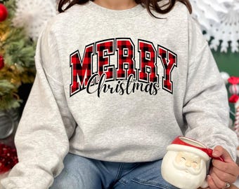 Puede incluir: Una sudadera gris claro con la inscripción "MERRY Christmas" en un diseño de cuadros rojos y negros. La sudadera se combina con unos vaqueros azules. Se sostiene una taza de Papá Noel.
