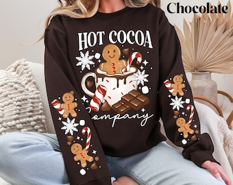 May include: Dark chocolate-colored sweatshirt with a festive "HOT COCOA Company" design. The graphic features a gingerbread man in a mug, candy canes, snowflakes, and chocolate bars. Gingerbread men and candy canes decorate the sleeves.