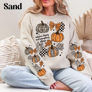 May include: A sand-colored sweatshirt featuring a fall-themed design. The design includes pumpkins, bows, and leaves in orange, black, and white. The text "autumn leaves and pumpkins please" is also visible.