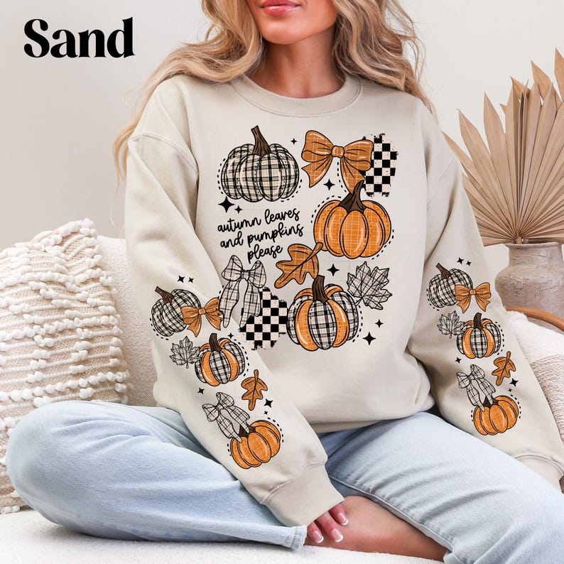 May include: A sand-colored sweatshirt featuring a fall-themed design. The design includes pumpkins, bows, and leaves in orange, black, and white. The text "autumn leaves and pumpkins please" is also visible.