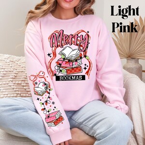May include: Light pink sweatshirt with the text "Merry Bookmas" in a festive design. The design includes stacks of books, string lights, and decorative elements. The sleeve features a matching design with books and lights.
