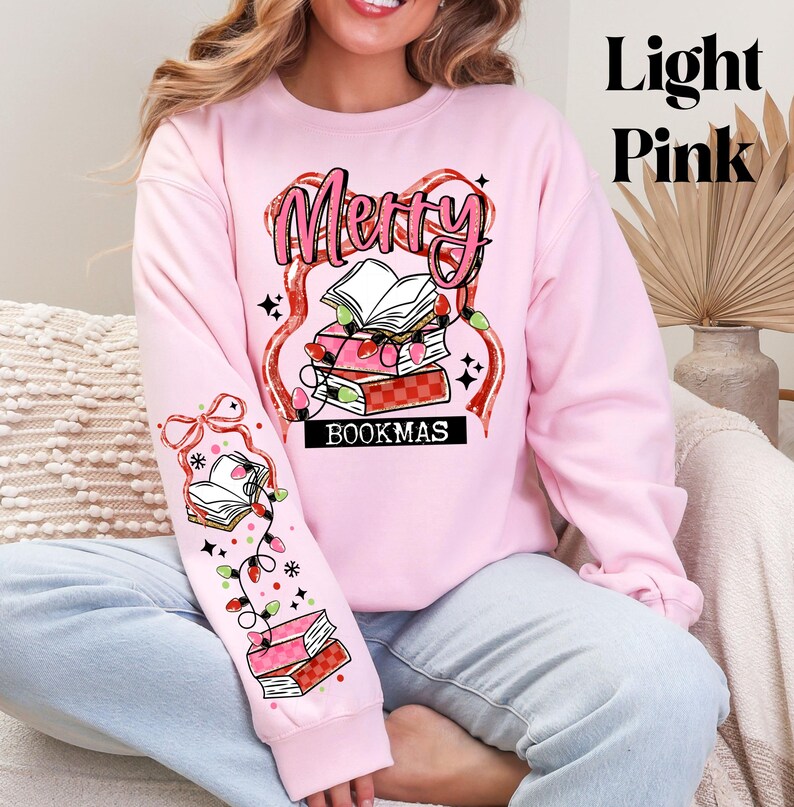 May include: Light pink sweatshirt with the text "Merry Bookmas" in a festive design. The design includes stacks of books, string lights, and decorative elements. The sleeve features a matching design with books and lights.