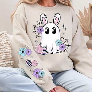 May include: Beige sweatshirt featuring a white ghost with bunny ears, black eyes, and purple and blue flowers. The ghost is surrounded by spiderwebs and Easter eggs. Additional flowers and eggs are on the sleeves.